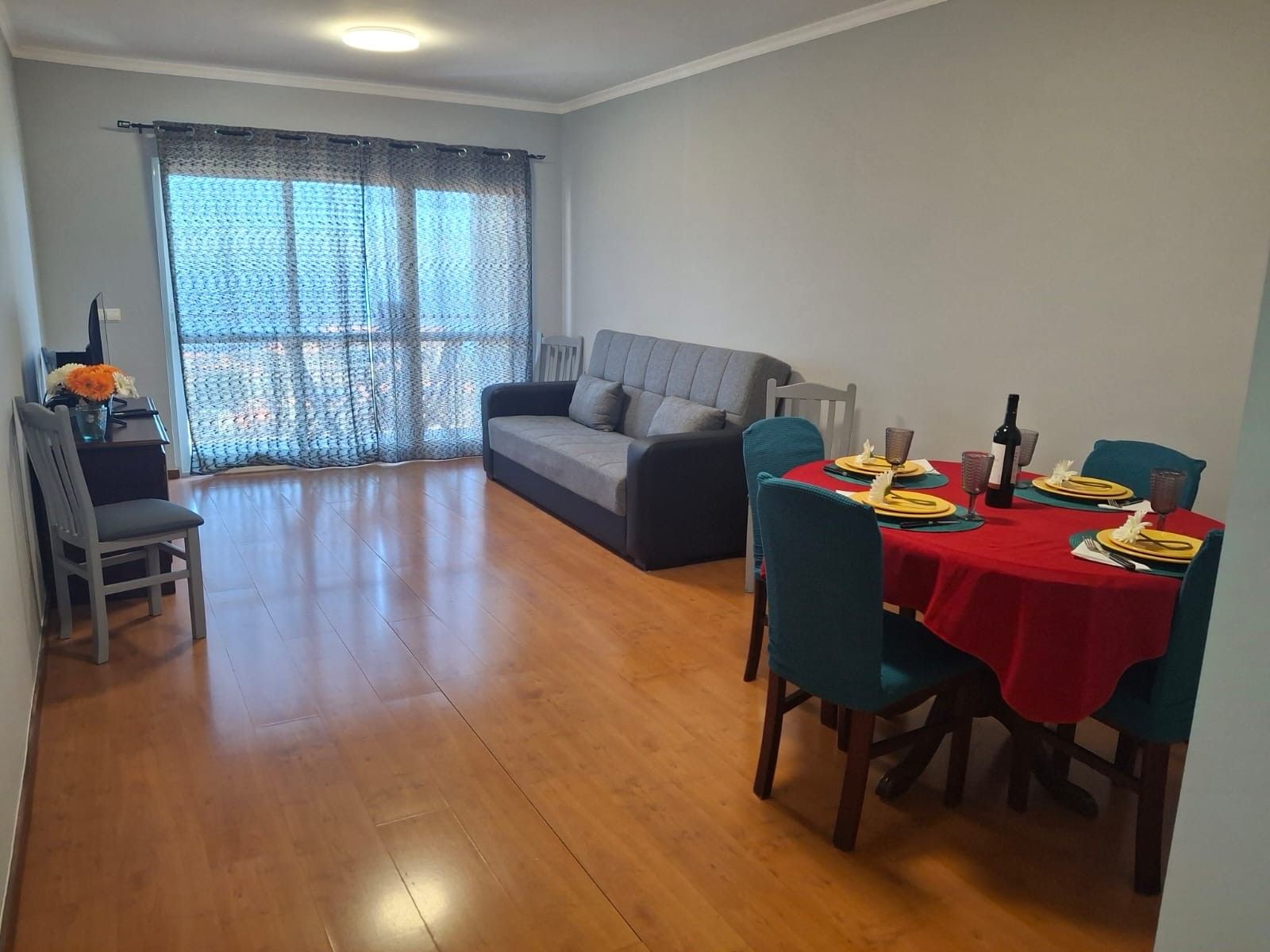 A dining area with a table set for four, a grey sofa, and a window with curtains.
