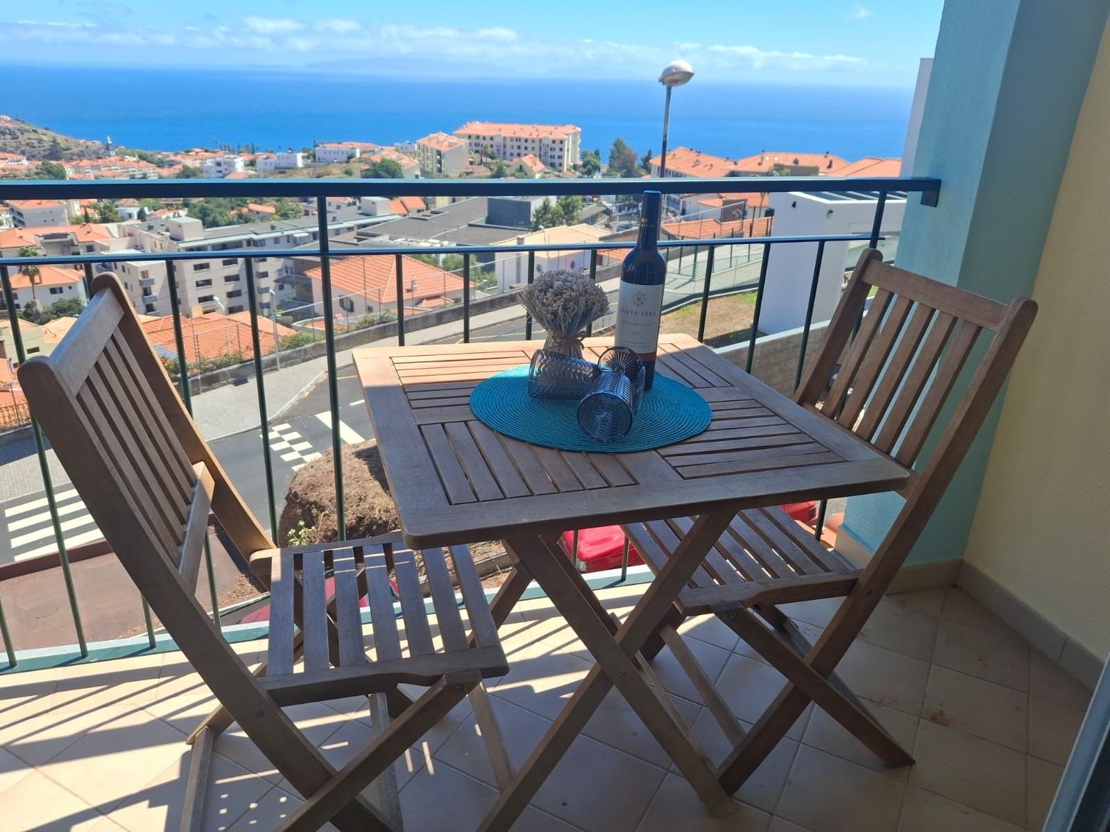 A balcony with a table and chairs, a bottle of wine, and a view of the sea and buildings.
