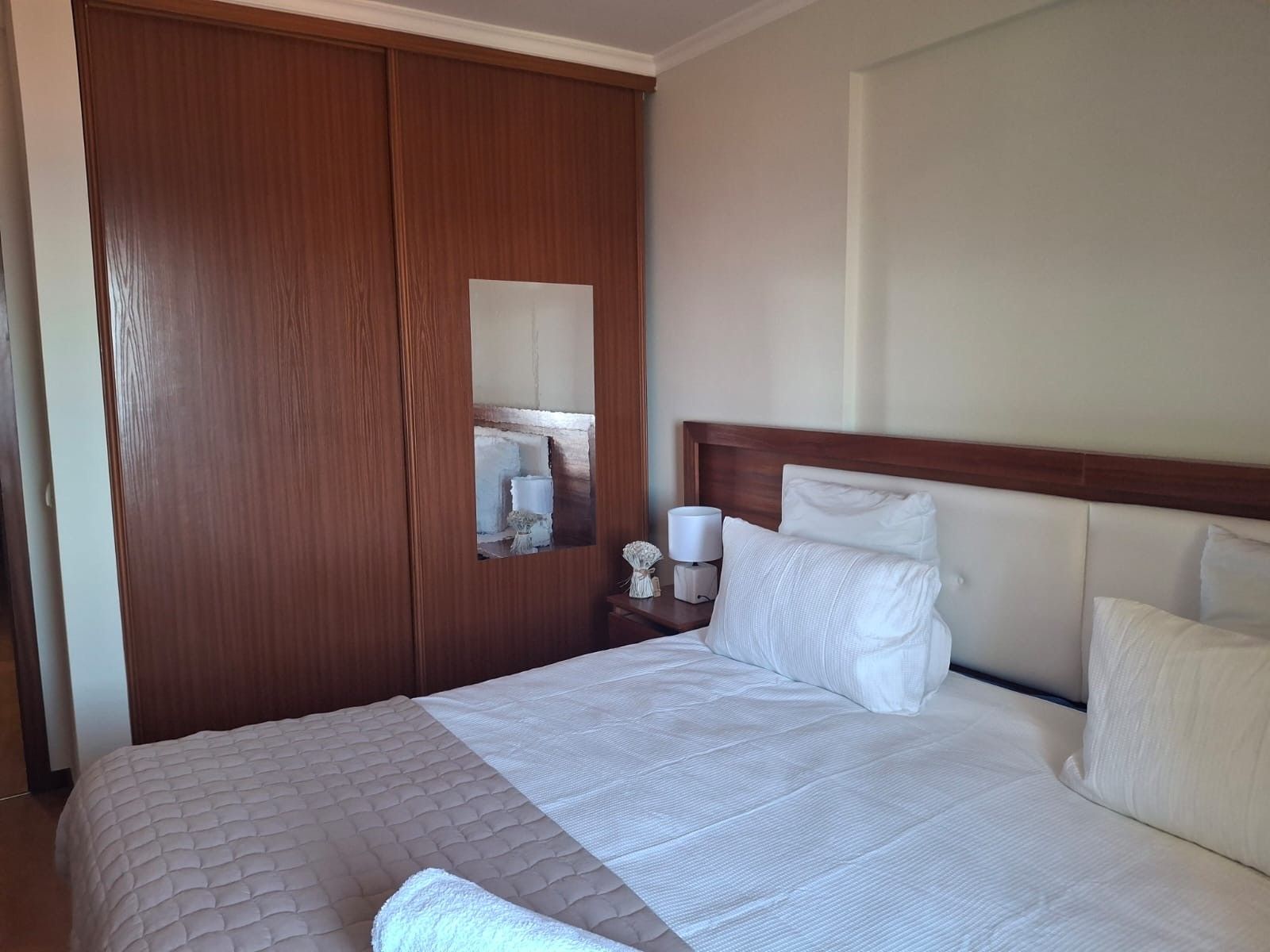 A hotel room with a large bed, a wooden wardrobe, a nightstand with a lamp, and a mirror.