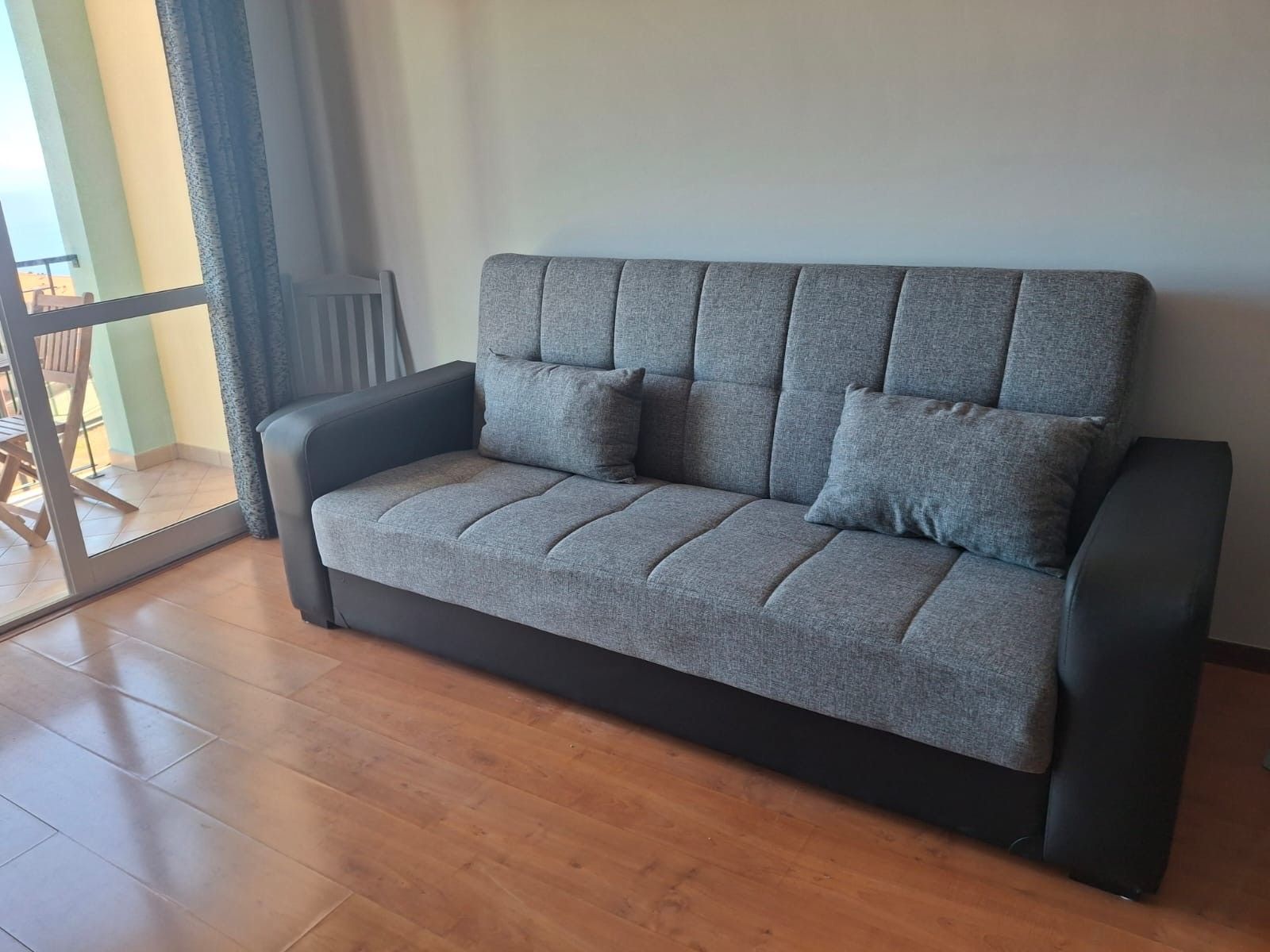 A grey sofa with a black frame in a room with wooden flooring and a sliding glass door leading to an outdoor area.