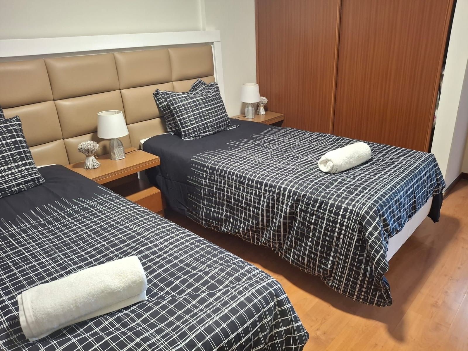 A hotel room with two beds, a wooden headboard, a nightstand with a lamp, and a wooden wardrobe.