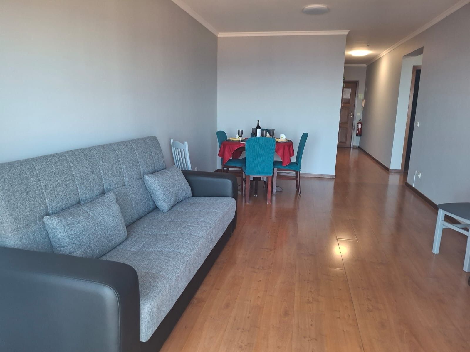 A modern living room with a grey sofa, a dining table with chairs, and a wooden floor.