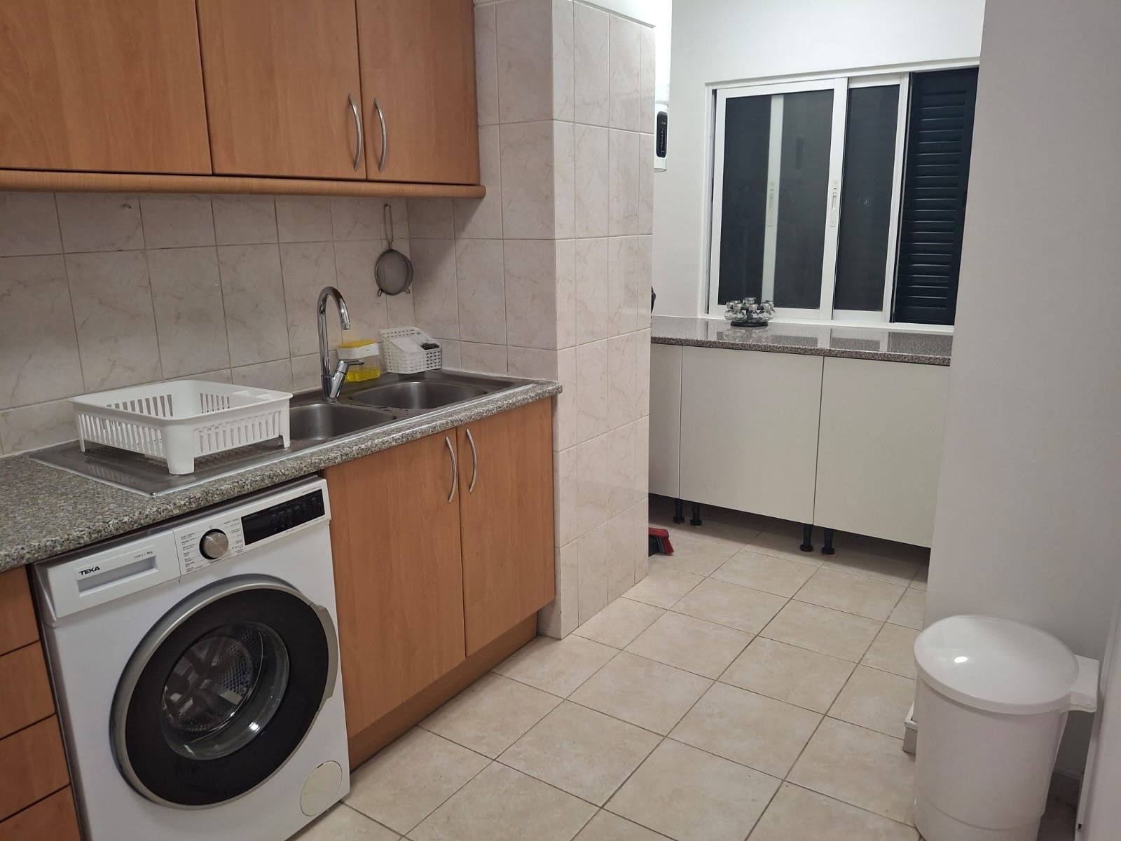 A compact kitchen with a washing machine, a sink, and a window with blinds.