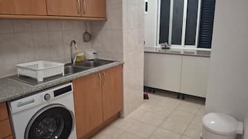 A compact kitchen with a washing machine, a sink, and a window with blinds.