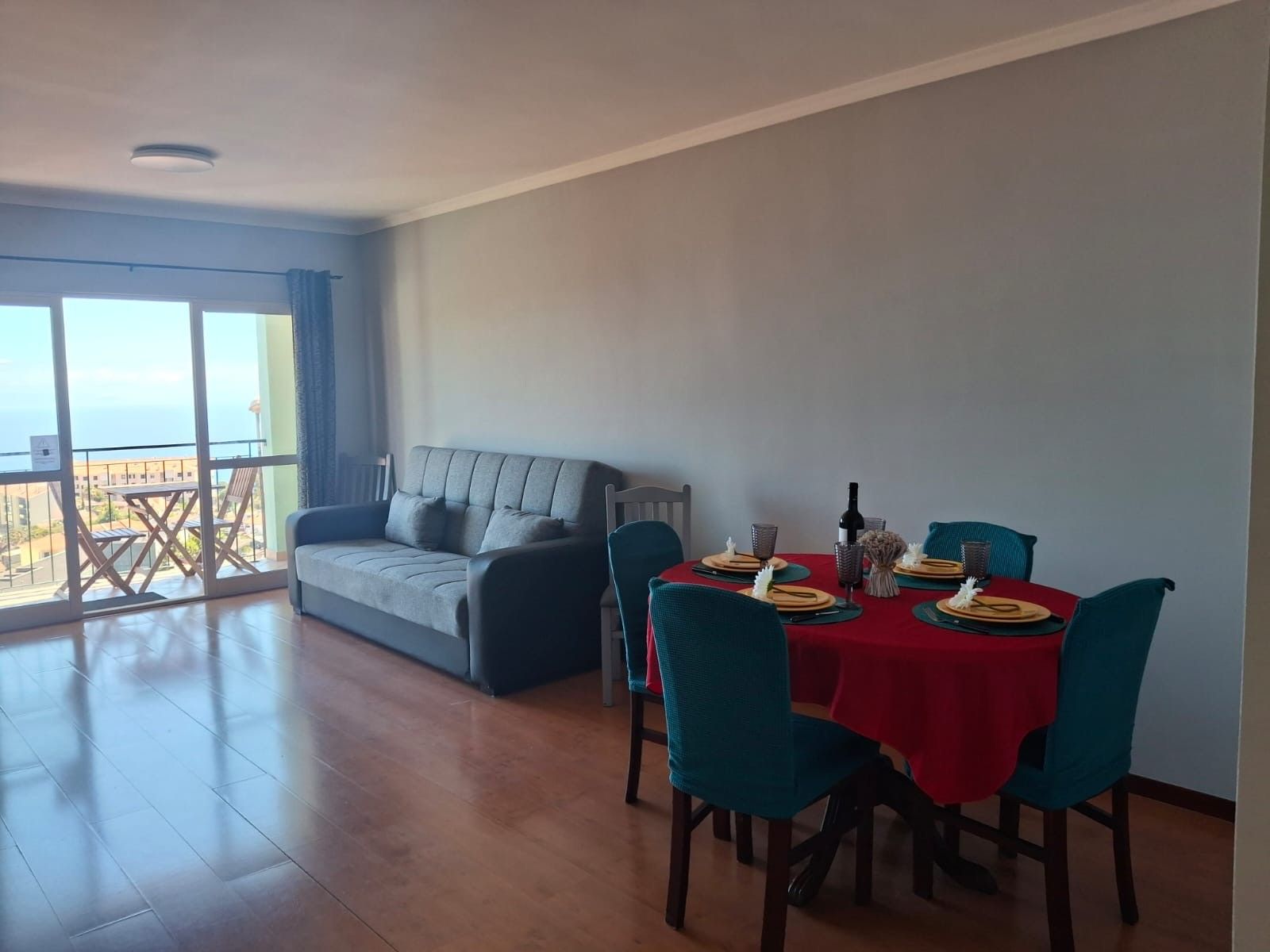 A dining area with a table set for four, a grey sofa, and a balcony with a view.