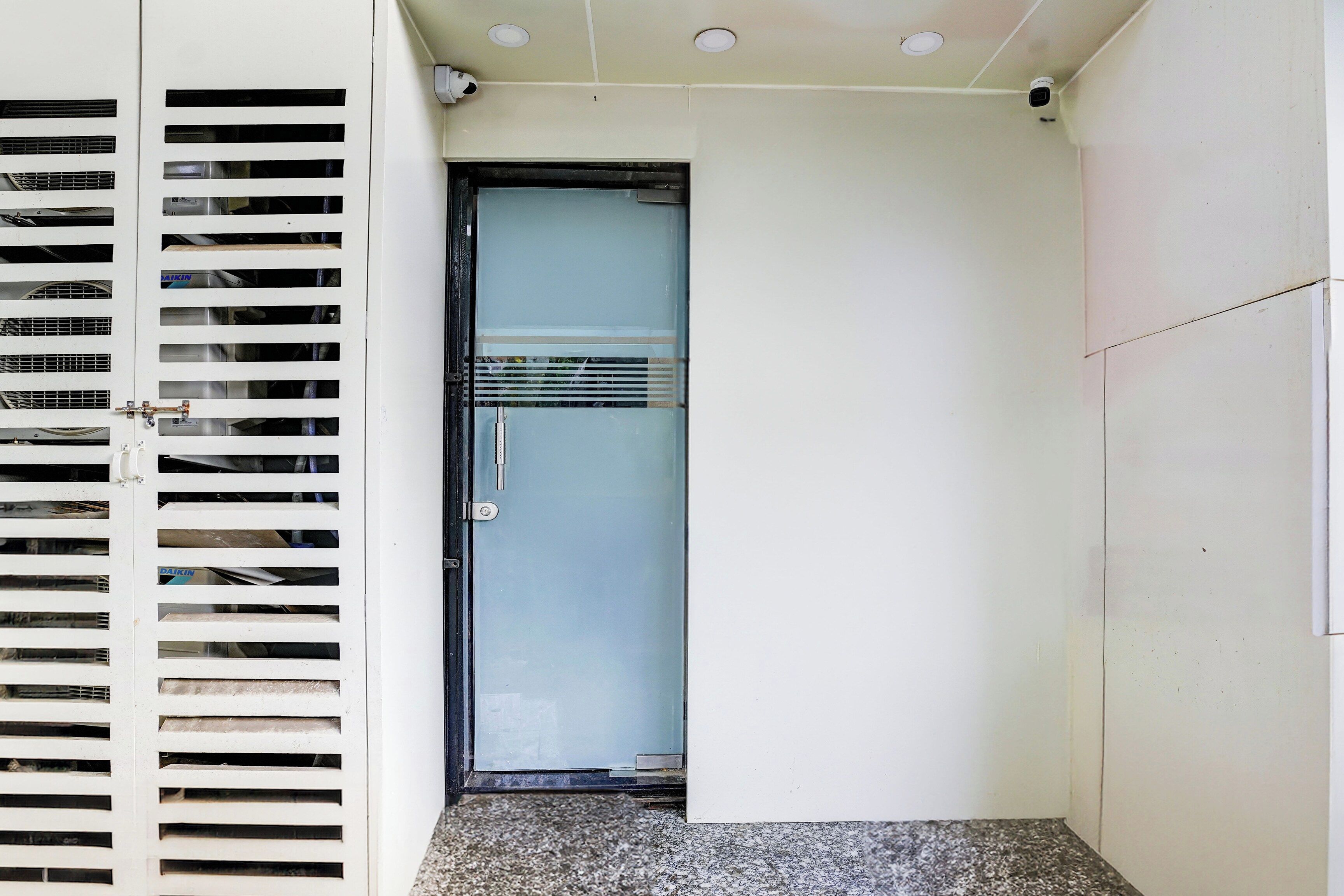Una puerta con manija metálica y un panel de vidrio, una pared blanca e iluminación empotrada en el techo.