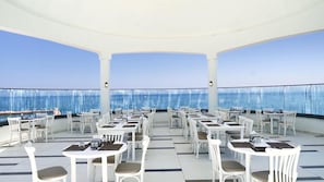 Ein Dachterrassen-Essbereich mit weißen Tischen und Stühlen, der Blick auf das Meer und Schiffe bietet.