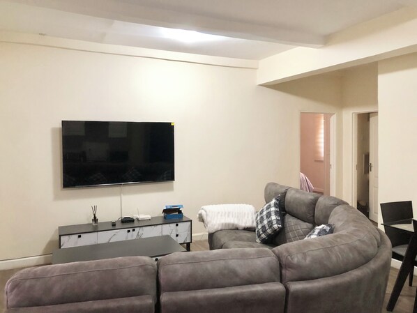 A living room with a large flat-screen TV mounted on the wall, a grey sectional sofa, a small side table, and a door leading to another room. - Apartment in secure complex near Pamodzi Hotel (Lusaka)