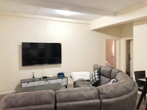 A living room with a large flat-screen TV mounted on the wall, a grey sectional sofa, a small side table, and a door leading to another room.