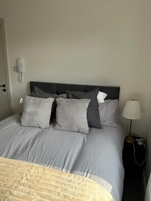 A neatly made bed with grey bedding, a bedside lamp, and a telephone on the wall. - Apartment Sleeps 2 With Shared Kitchen Near City (London)