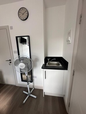 A compact kitchen with a sink, a clock, and a standing fan. - Apartment Sleeps 2 With Shared Kitchen Near City (London)