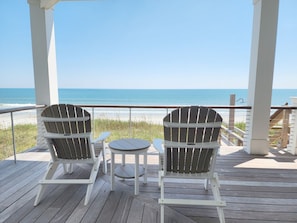 Eine überdachte Terrasse mit zwei Adirondack-Stühlen und einem kleinen Tisch, mit Blick auf einen Strand.