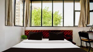Quarto de hotel com uma cama grande, cabeceira estofada em vermelho, escrivaninha com cadeira e janela com cortinas.