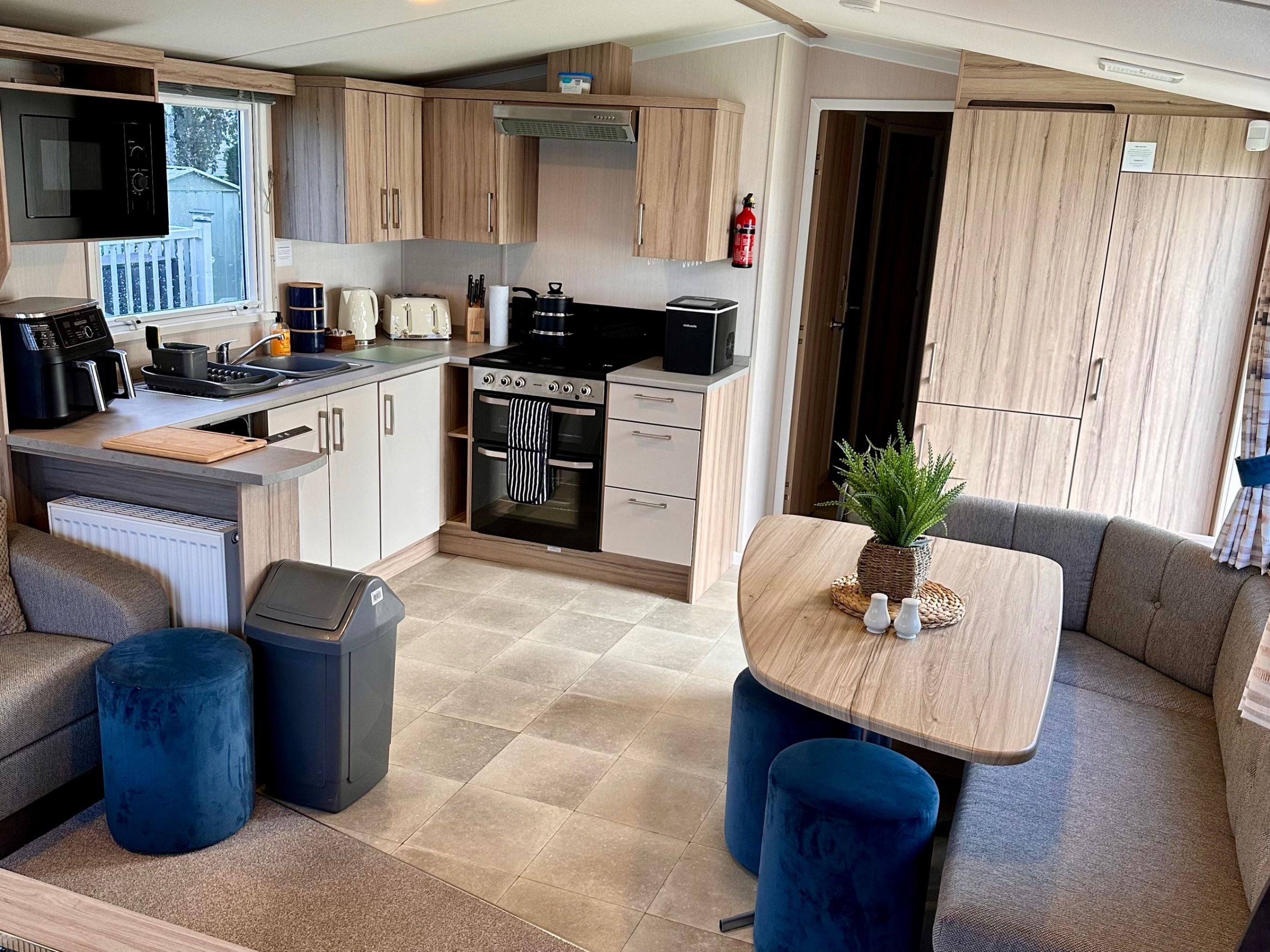 Een compact keukenhoekje met een fornuis, oven en magnetron, een zithoek met een tafel en blauwe poefs, en uitzicht naar buiten door een raam.