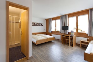 Habitación de hotel con puerta de madera, dos camas, un escritorio con silla, televisor y ventanas amplias con cortinas.
