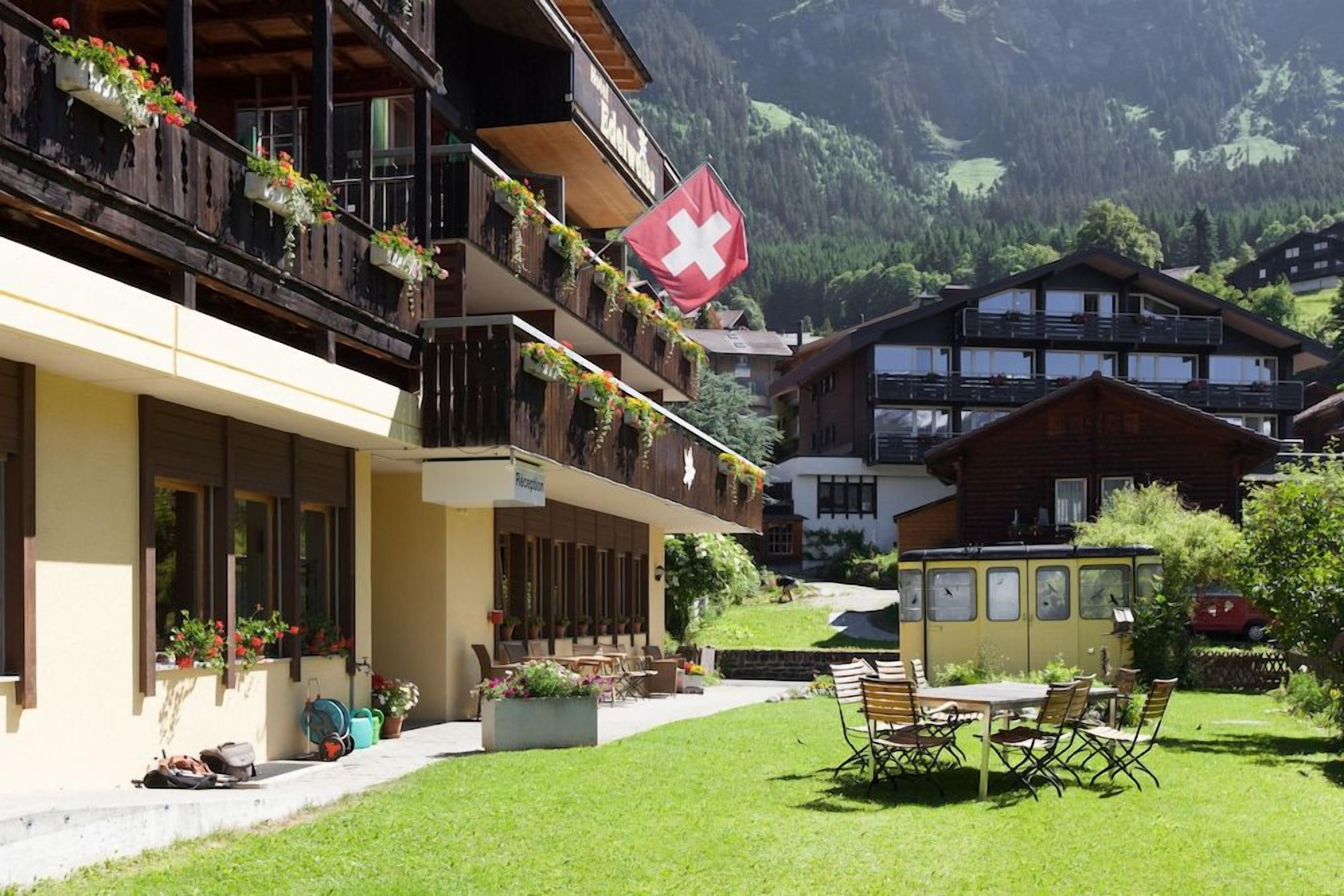A hotel with balconies adorned with flowers, a Swiss flag, and a view of mountains.