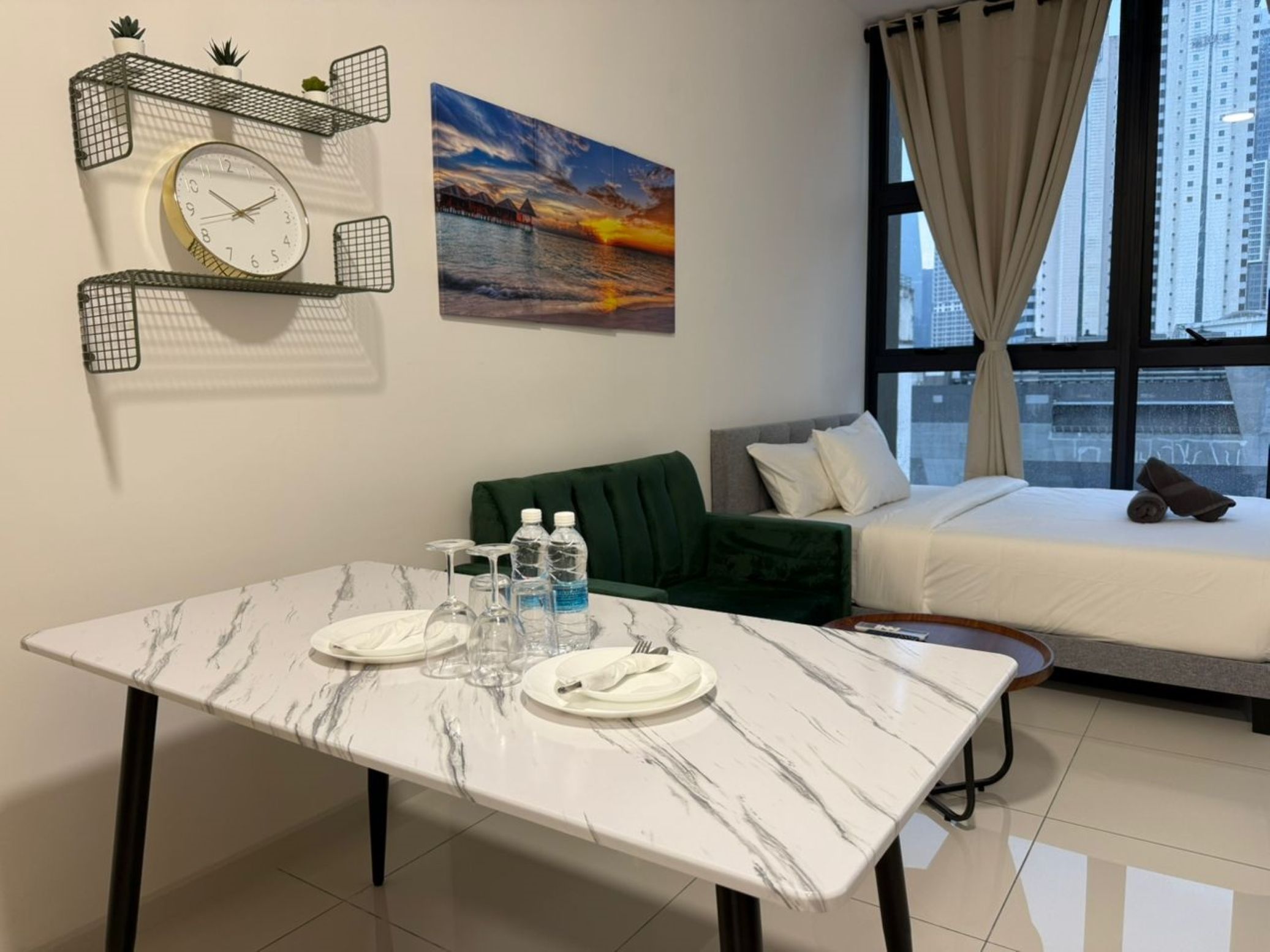 Habitación de hotel con una cama, un sofá verde, una mesa de mármol preparada para dos personas, un reloj de pared y un cuadro con una vista al atardecer.