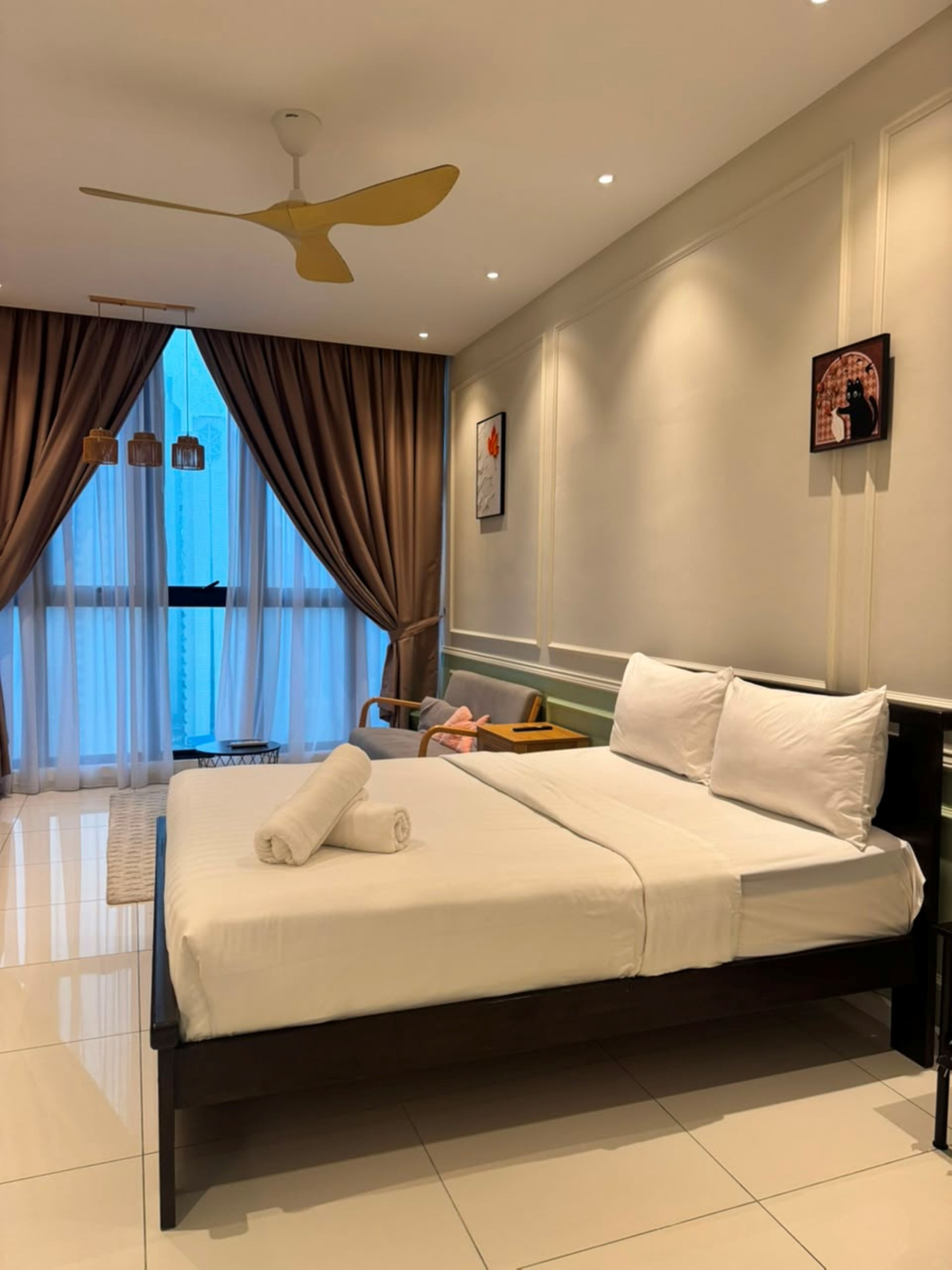Una habitación de hotel con una cama grande, un ventilador de techo, una ventana con cortinas y un cuadro en la pared.