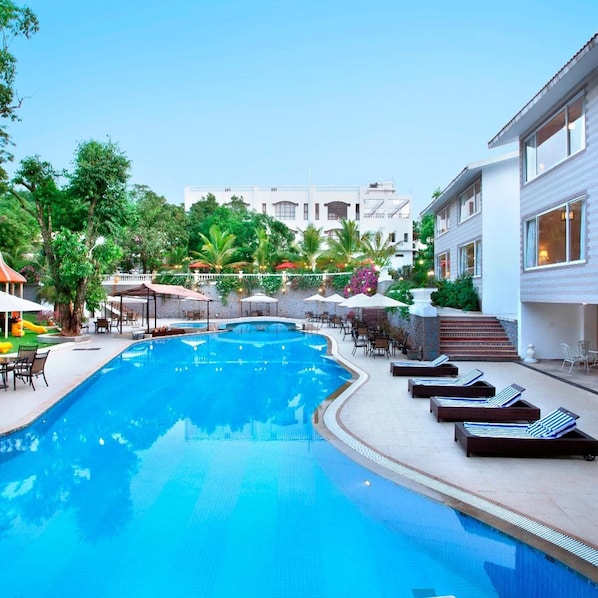 Une piscine avec des chaises longues, un espace de détente couvert et un bâtiment en arrière-plan.