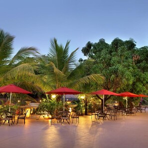 An outdoor seating area with tables and chairs under red umbrellas, surrounded by palm trees and other greenery. - Villa San Lawrenz (Mawal)