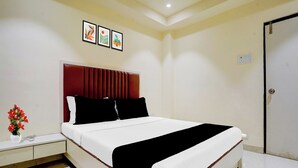 A hotel room with a bed, a nightstand, a vase with flowers, and a door. - O Ulhasnagar Formerly Hotel Derby (Mumbai)