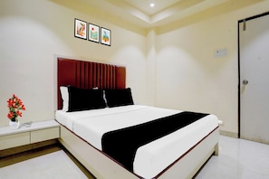 A hotel room with a bed, a nightstand, a vase with flowers, and a door. - O Ulhasnagar Formerly Hotel Derby (Mumbai)