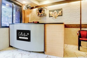 A reception desk with a sign indicating it's a company-serviced collection point, a wall-mounted fan, and a framed picture above the desk. - O Ulhasnagar Formerly Hotel Derby (Mumbai)