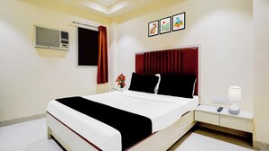 A hotel room with a bed, a bedside table with a lamp, a wall-mounted air conditioner, and a window with a curtain. - O Ulhasnagar Formerly Hotel Derby (Mumbai)