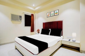 A hotel room with a bed, a bedside table with a lamp, a wall-mounted air conditioner, and a window with a curtain. - O Ulhasnagar Formerly Hotel Derby (Mumbai)