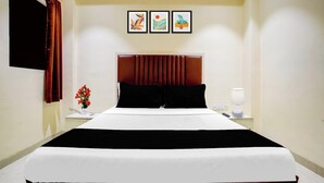 A hotel room with a large bed, a nightstand with a lamp, a vase with flowers, and three framed pictures on the wall. - O Ulhasnagar Formerly Hotel Derby (Mumbai)