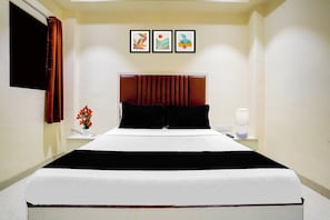 A hotel room with a large bed, a nightstand with a lamp, a vase with flowers, and three framed pictures on the wall. - O Ulhasnagar Formerly Hotel Derby (Mumbai)