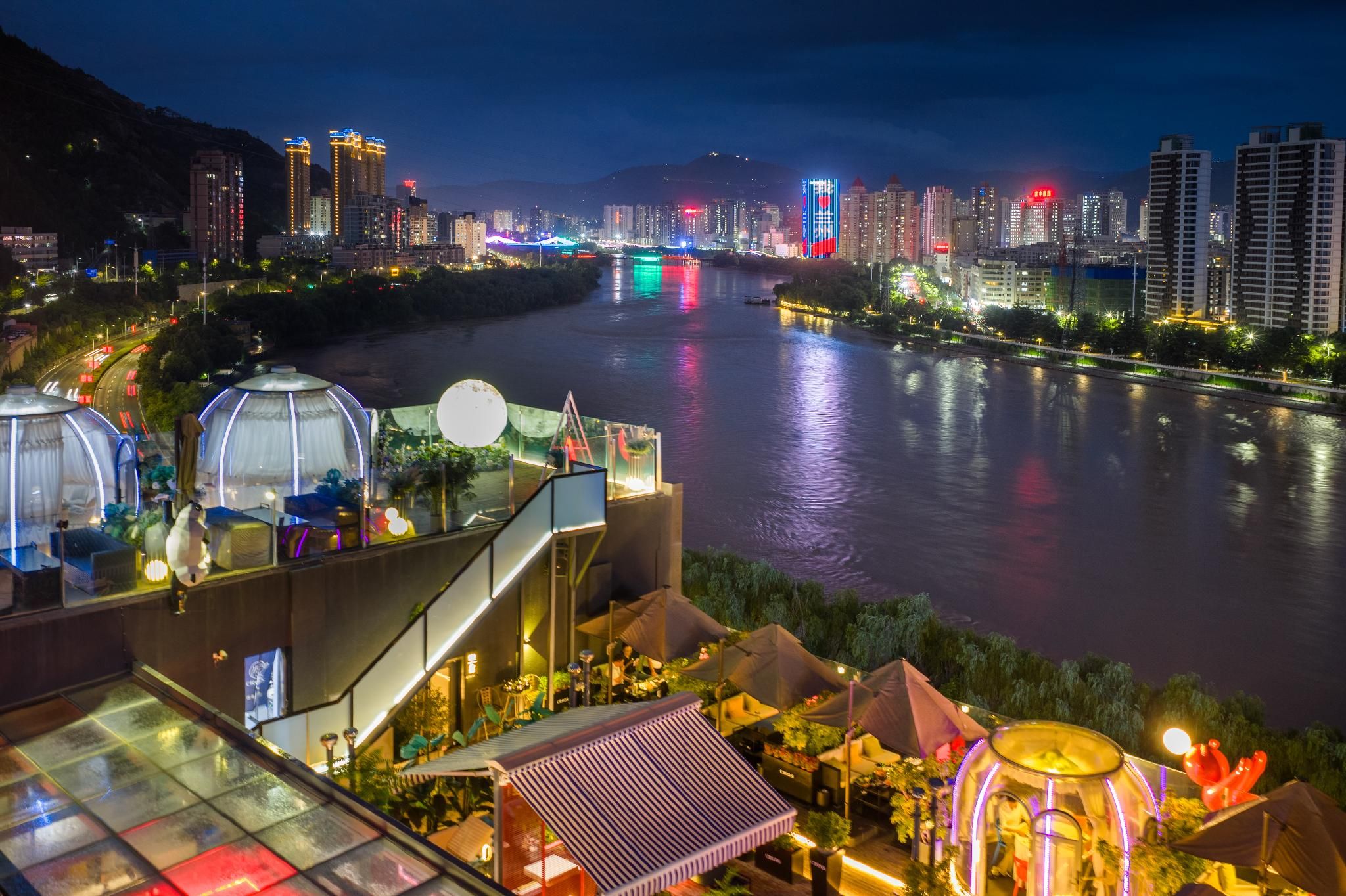 A riverside view of a hotel with a glass-roofed terrace, outdoor seating, and a city skyline in the background.