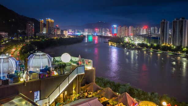 A riverside view of a hotel with a glass-roofed terrace, outdoor seating, and a city skyline in the background. - Atour Hotel (Lanzhou)