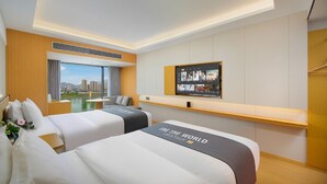 Habitación de hotel con dos camas, un ventanal con vistas a la ciudad, un televisor de pantalla plana en la pared y una mesita con una lámpara.