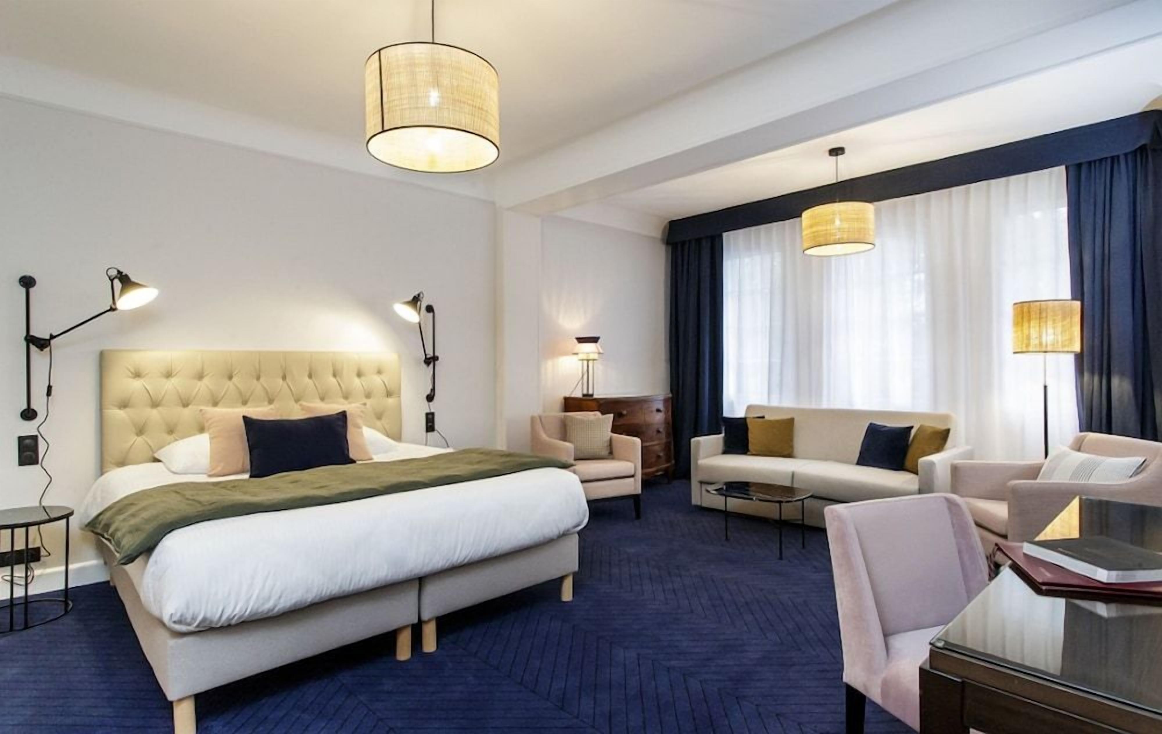 Une chambre d’hôtel avec un grand lit, un canapé, un fauteuil, un bureau et deux lampes suspendues.
