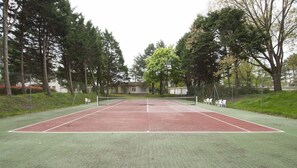 Una cancha de tenis con red, rodeada de árboles y un edificio al fondo.