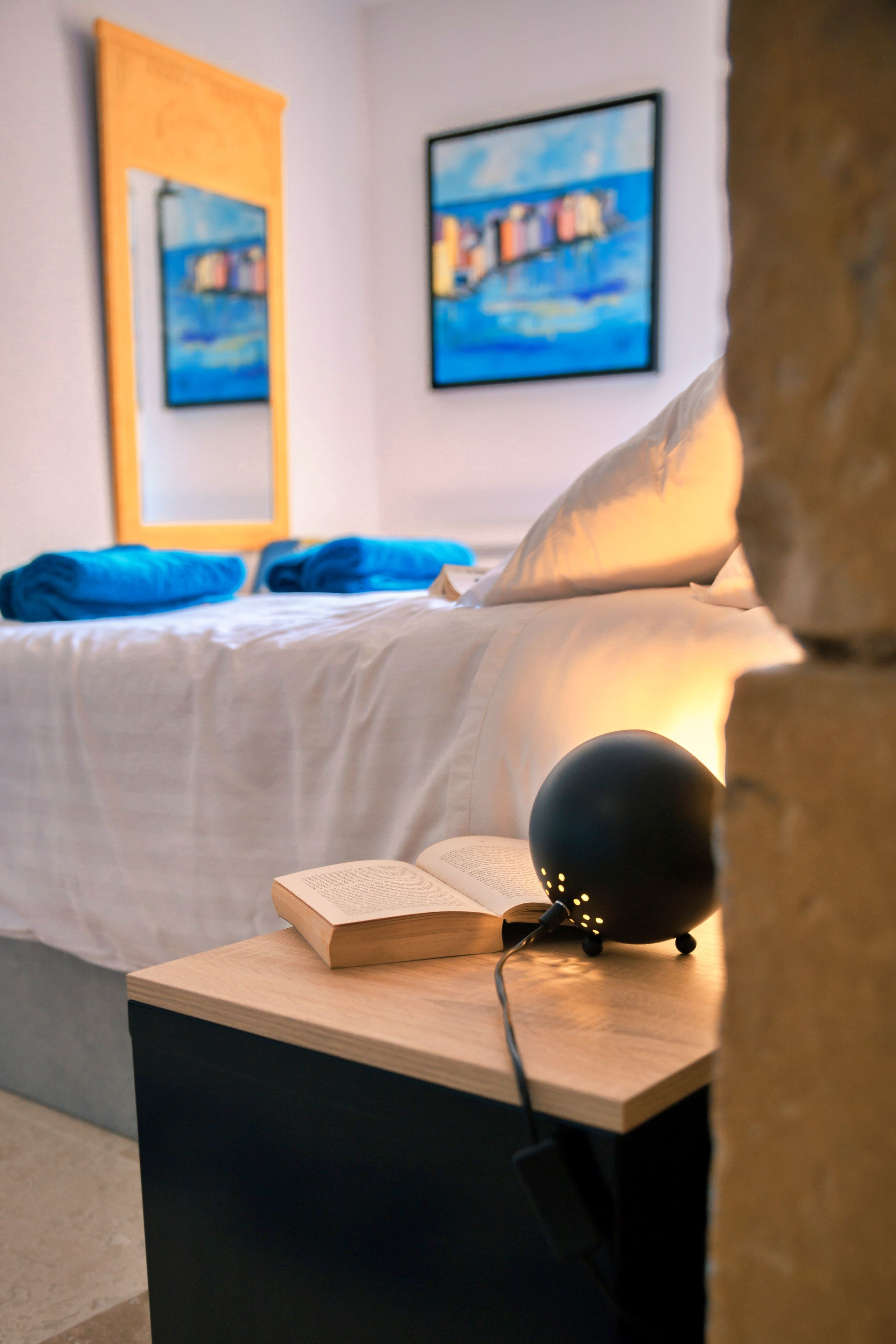A bedroom with a bed, a bedside table with a book and a lamp, and two paintings on the wall.