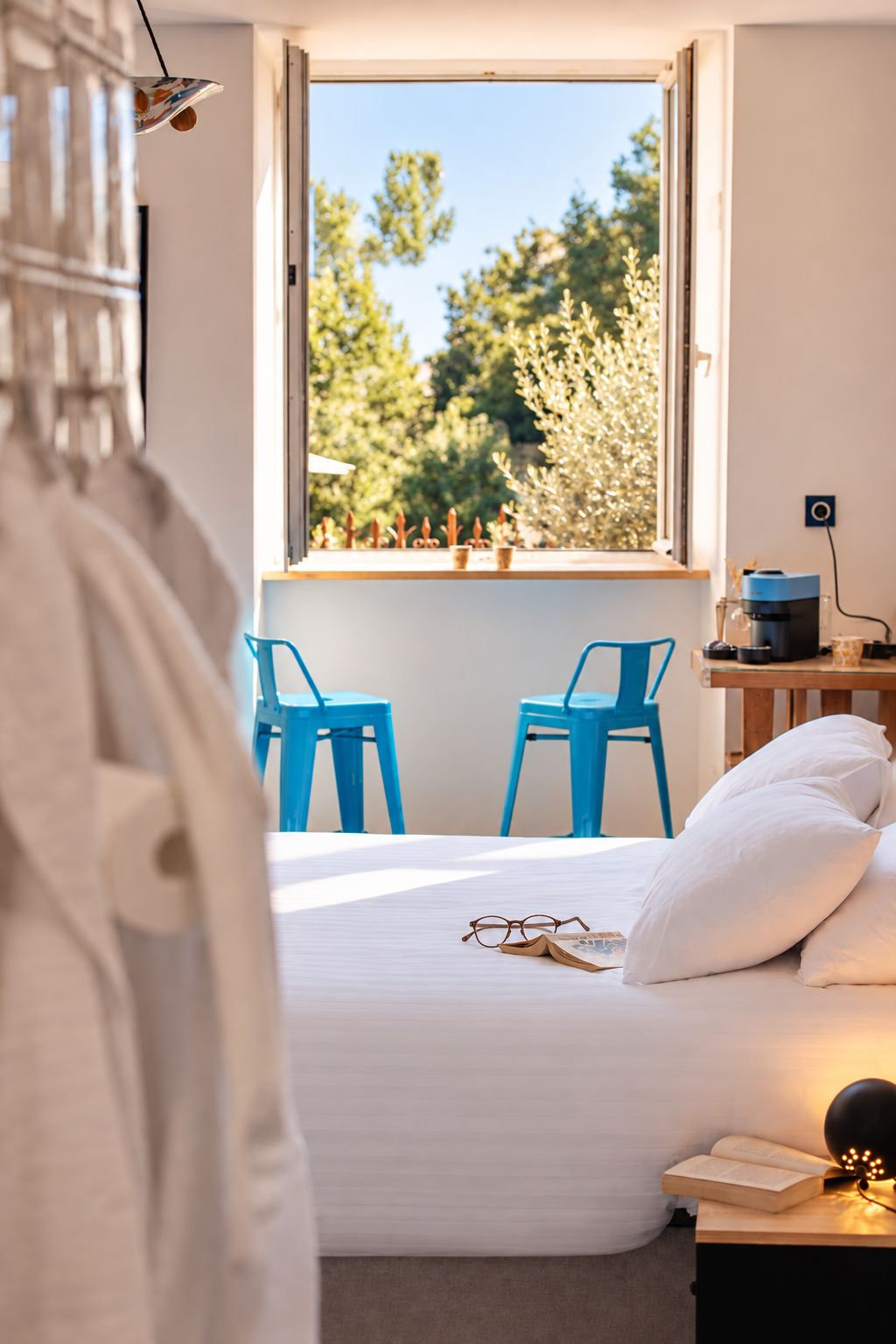 A bedroom with a bed, two blue chairs, a window with a view of trees, and a bedside table with a lamp and a book.