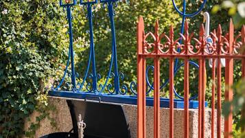 Una griglia da barbecue con un recinto decorativo blu e rosso, un lampione e vegetazione lussureggiante sullo sfondo.