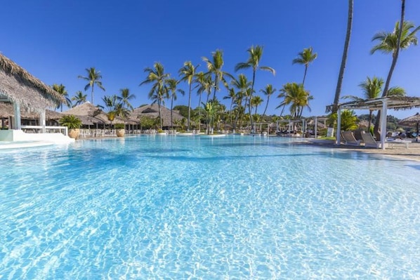 Una piscina azul cristalina rodeada de palmeras y construcciones con techo de paja.