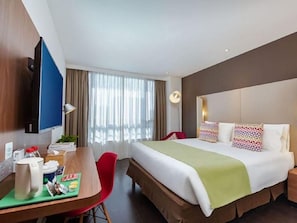Une chambre d’hôtel équipée d’un lit, d’une table de chevet, d’un bureau, d’une chaise et d’un téléviseur fixé au mur.