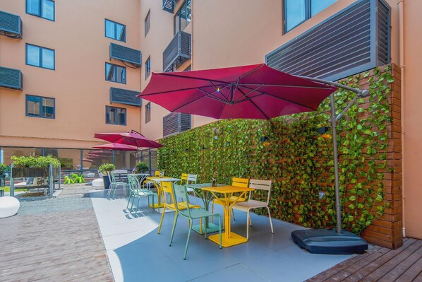 An outdoor seating area with tables and chairs, a red umbrella, and a wall covered with green foliage.