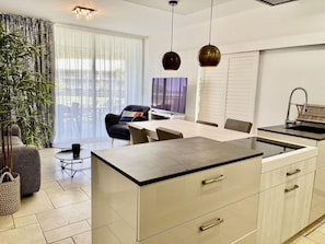 A modern kitchen with a central island, a dining area with chairs, a television, and a large window. - Anse Marcel Beach Resort (Anse Marcel)