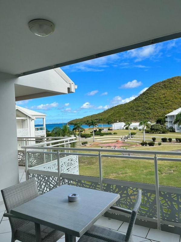 A balcony with a table and chairs, overlooking a coastal area with buildings and a hill. - Anse Marcel Beach Resort (Anse Marcel)