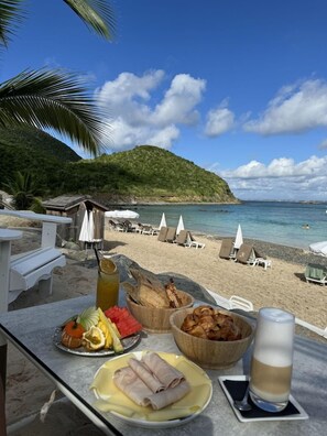 A beachside dining area with a view of the sea, a wooden table with food and drinks, and beach chairs in the background. - Anse Marcel Beach Resort (Anse Marcel)