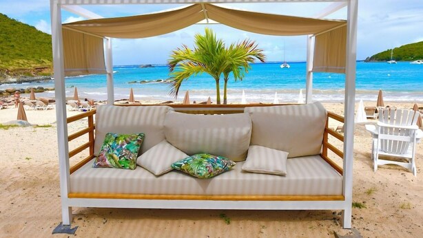 Una cama junto a la playa con dosel, cojines blancos y una almohada tropical.