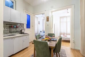 Una cucina compatta con mobili bianchi, una zona pranzo con tavolo apparecchiato per due e vista su una camera da letto con letto e tende.