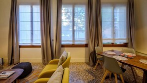 Ein Zimmer mit Esstisch, Stühlen, einer Couch und großen Fenstern mit Vorhängen.
