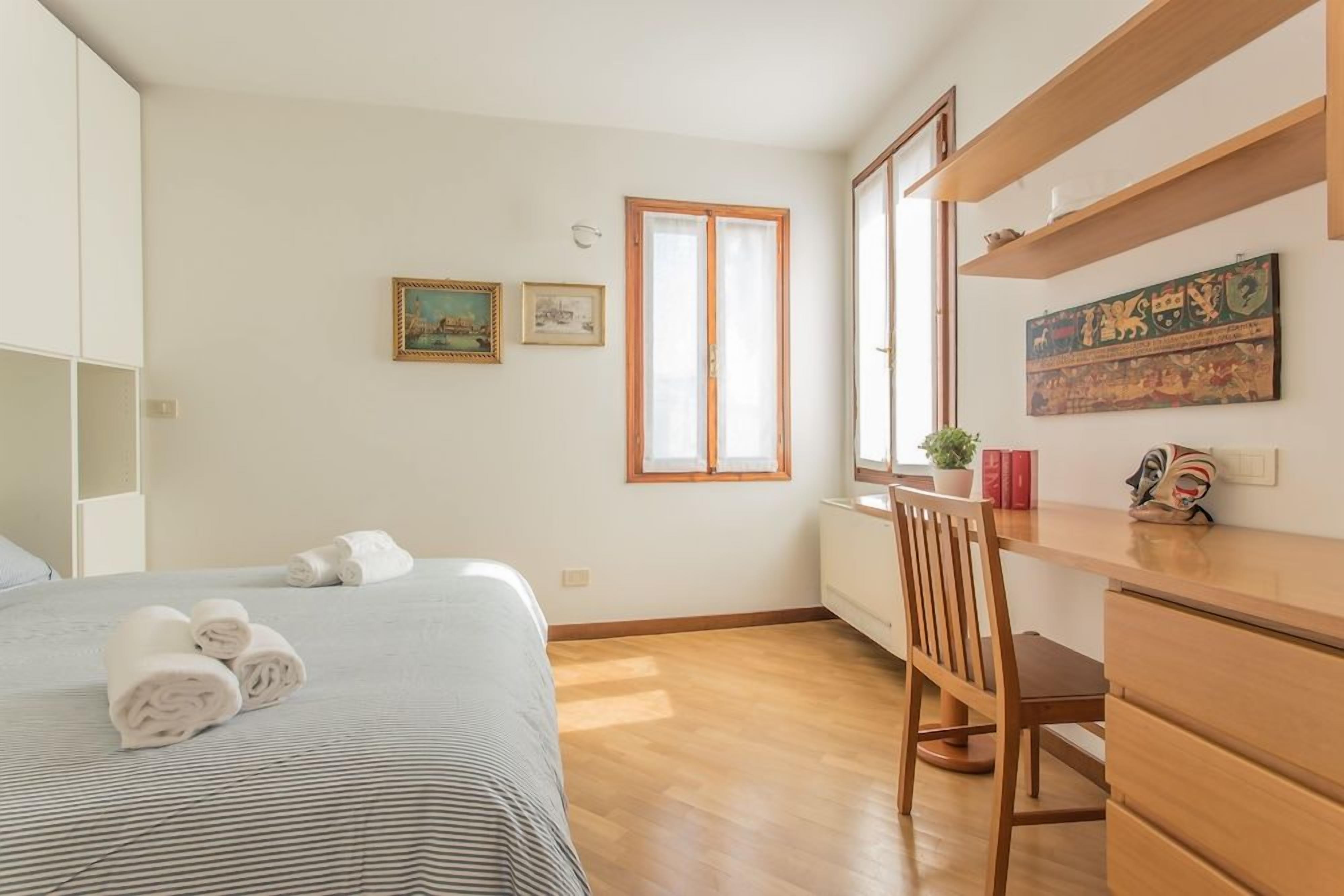 Un dormitorio con cama, escritorio y silla. Hay dos ventanas, una con vista al exterior y otra al interior.