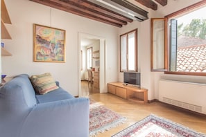 A living room with a blue sofa, a television, a wooden cabinet, and a large window with a view of a tiled roof. - Venezia - Castello 1542 (Venice)